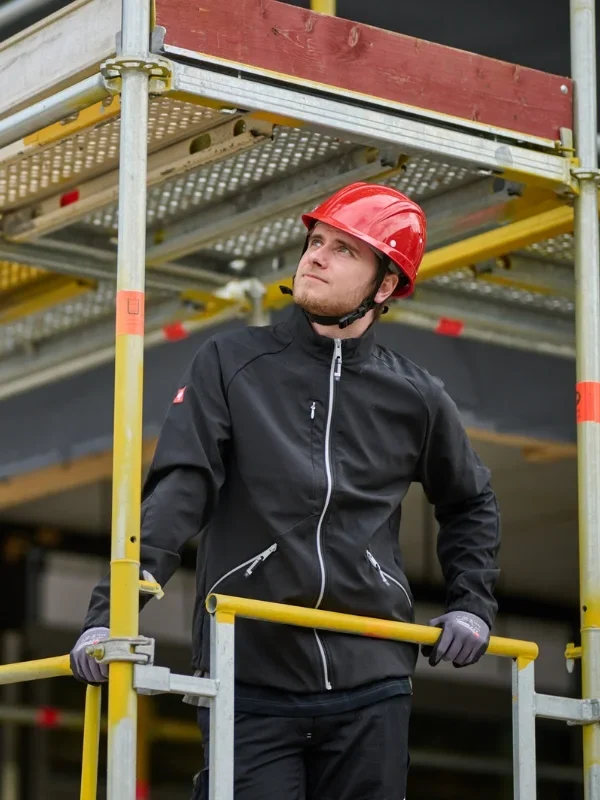 Ein Bauarbeiter mit rotem Schutzhelm steht auf einem Gerüst und schaut nach oben, während er sich an den Geländern festhält.